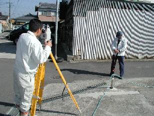 地籍測量を行っている写真。黄色い三脚の器具を用い、作業員2人が測量を行っている。