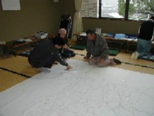 地図と簿冊の閲覧のようす。和室一杯に大きく地図が広げられ、関係者3名で確認を行っている。