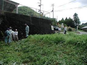 所有者、地番、地目、境界の調査を行っている写真。雑草の生えた土地の上に緑のツナギを着た作業員、及び関係者ら6人ほどがいる。